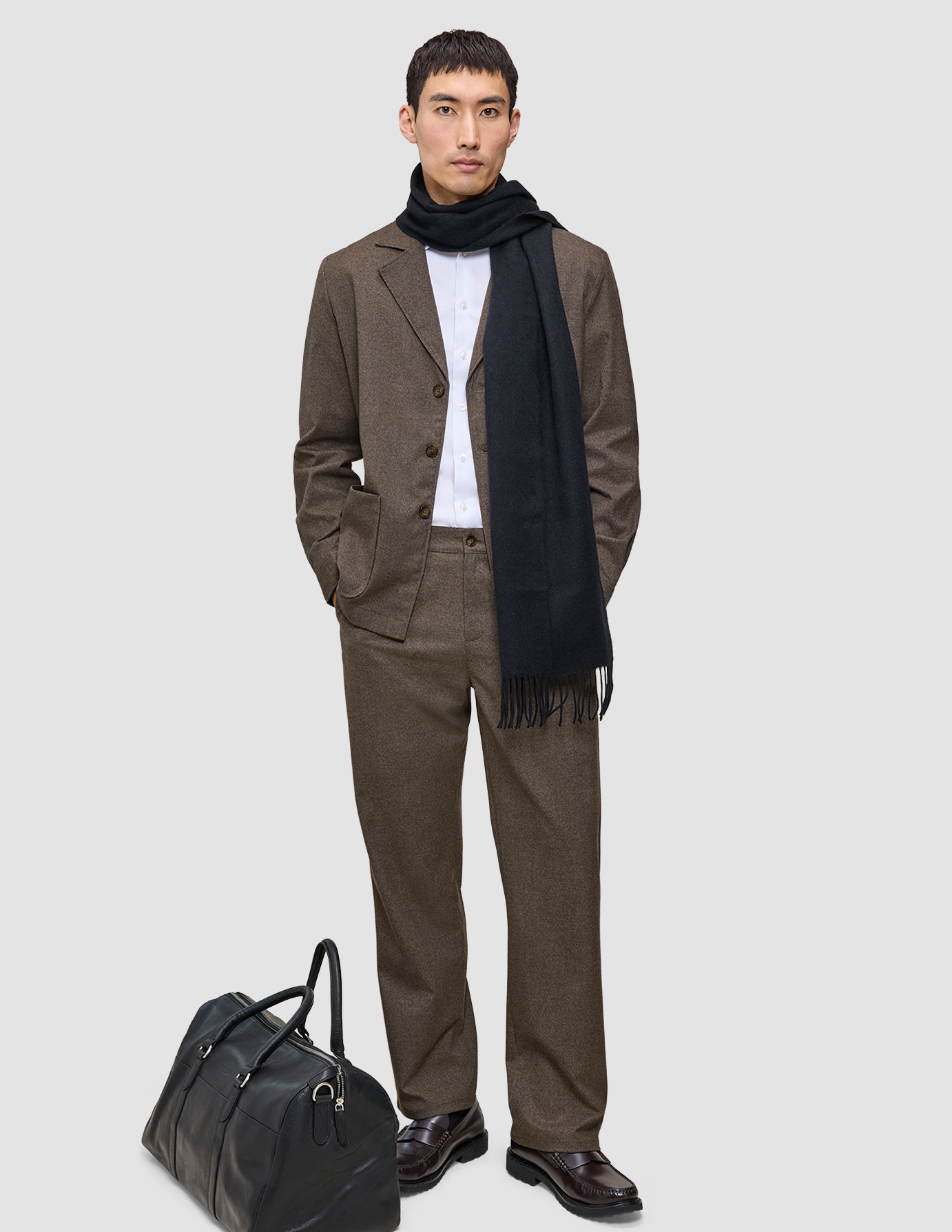 En mand iført en brun Atelier Overshirt Dark Beige, en hvid skjorte og et sort tørklæde står ved siden af en sort læderrejsetaske.